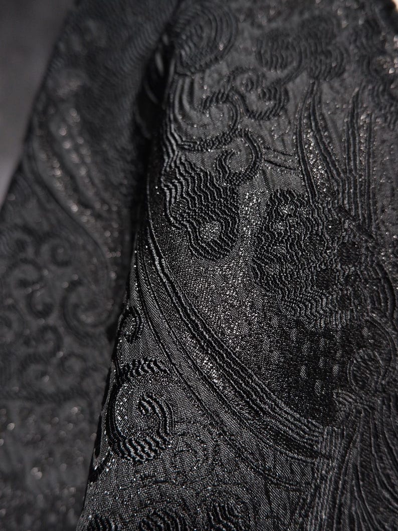 K&ouml;nnte beinhalten: Nahaufnahme eines schwarzen Stoffes mit einem komplizierten, erhabenen Paisley-Design. Der Stoff hat einen subtilen Glanz, mit einer strukturierten Oberfl&auml;che und detaillierten Mustern. Das Design ist ein sich wiederholendes Motiv, das einen luxuri&ouml;sen Look erzeugt.