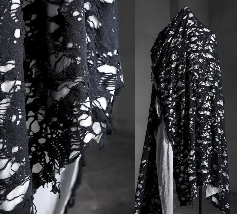 May include: Black fabric with a distressed, ripped, and torn design. The fabric has a lace-like appearance with large holes throughout. The fabric is draped over a mannequin.