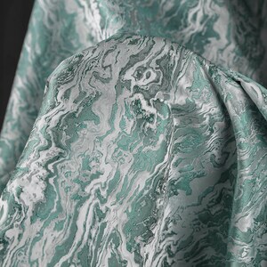 K&ouml;nnte beinhalten: Nahaufnahme eines teal- und wei&szlig;en Stoffes mit Marmormuster. Der Stoff ist weich und hat einen dezenten Glanz.