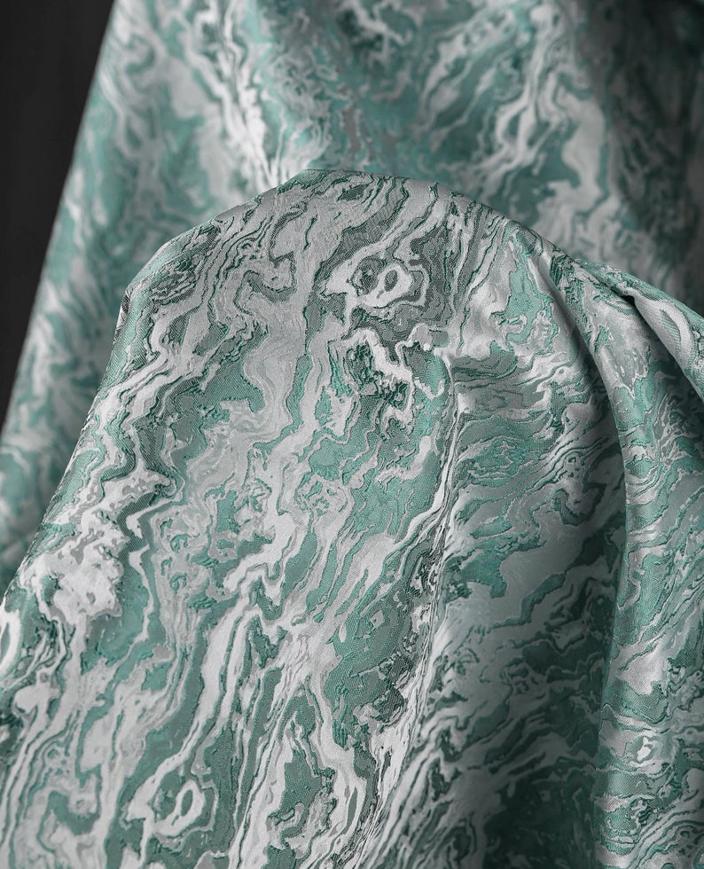 K&ouml;nnte beinhalten: Nahaufnahme eines teal- und wei&szlig;en Stoffes mit Marmormuster. Der Stoff ist weich und hat einen dezenten Glanz.