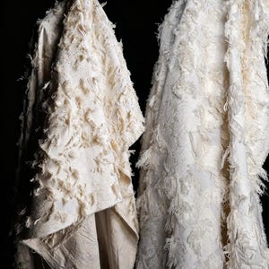 May include: Two pieces of off-white fabric featuring a textured, frayed fringe design. The material has a layered, shaggy appearance with loose threads and a woven base, creating a dimensional surface against a solid black background.