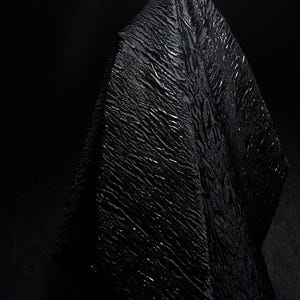 Op de afbeelding: Een close-up van een zwarte, getextureerde stof met een gerimpeld oppervlak, tegen een donkere achtergrond. De textuur van de stof vangt het licht op en creëert highlights. De afbeelding heeft een dramatische en mysterieuze uitstraling.