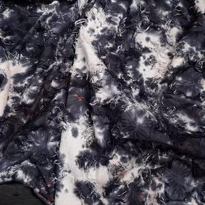 May include: Close-up of a fabric with a tie-dye pattern in shades of black, white, and gray. The fabric has a textured, fringed appearance, with frayed edges and a soft, flowing drape. The design features irregular, blotchy patterns.
