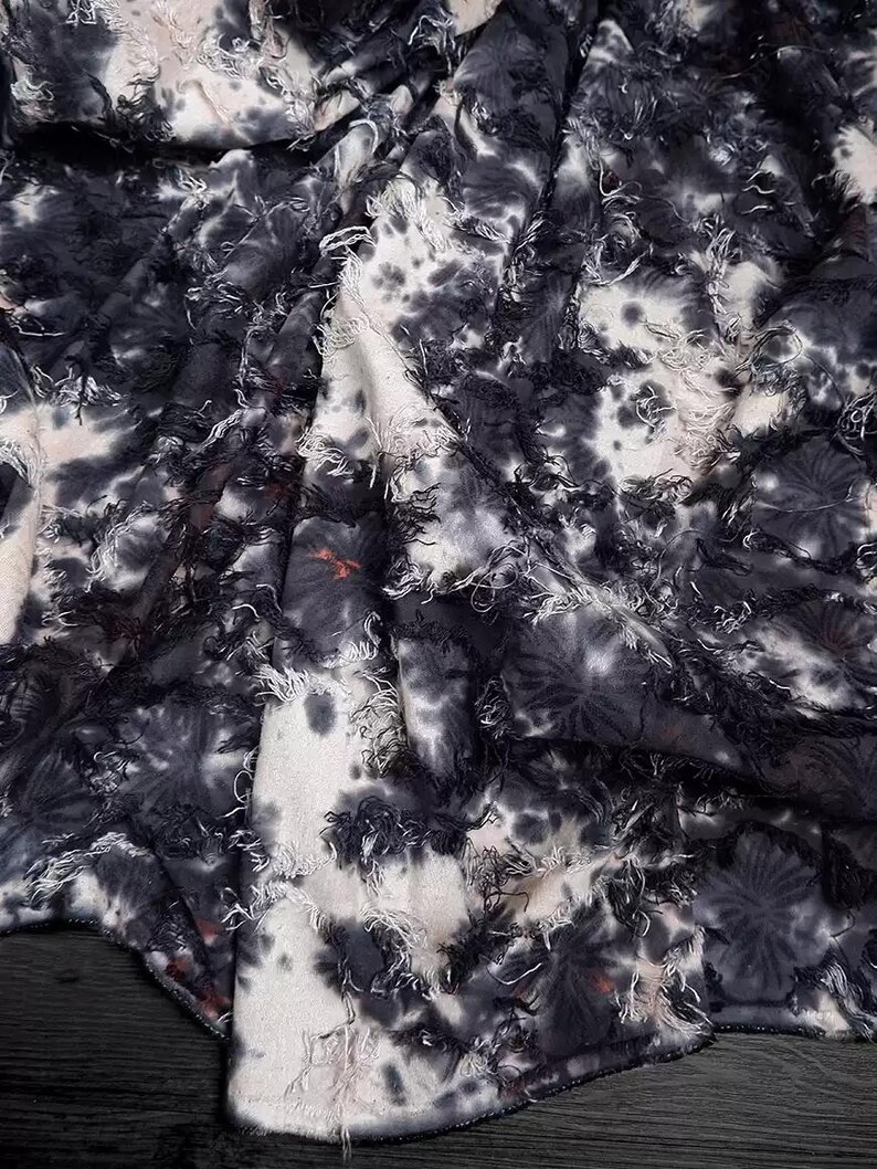 May include: Close-up of a fabric with a tie-dye pattern in shades of black, white, and gray. The fabric has a textured, fringed appearance, with frayed edges and a soft, flowing drape. The design features irregular, blotchy patterns.