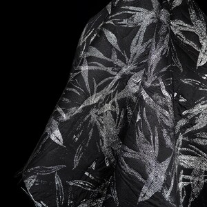 Puede incluir: Tela negra con un diseño de hojas plateadas. La tela es texturizada y parece estar drapeada, con las hojas plateadas creando un efecto brillante sobre el fondo oscuro. El aspecto general es elegante y sofisticado.
