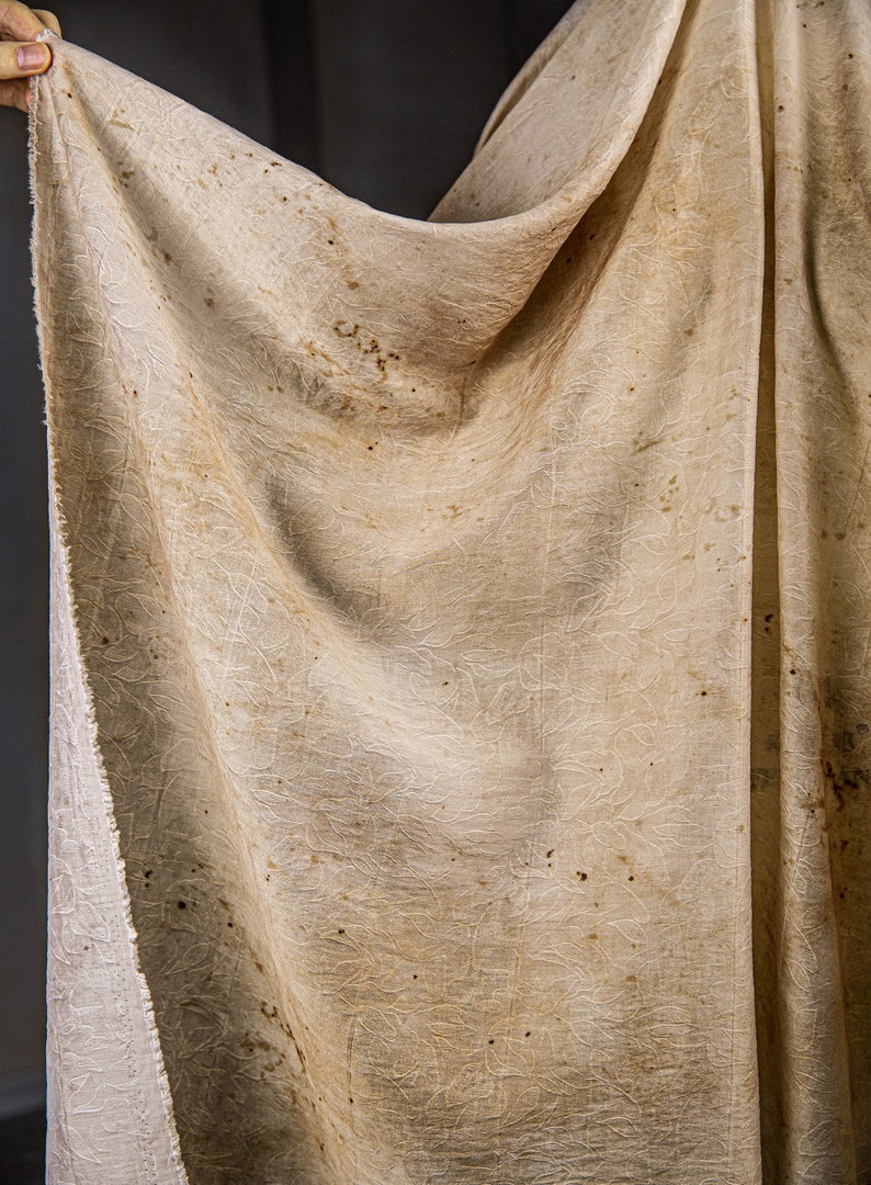 May include: A beige fabric with a textured pattern and brown spots. The fabric is draped over a hand.