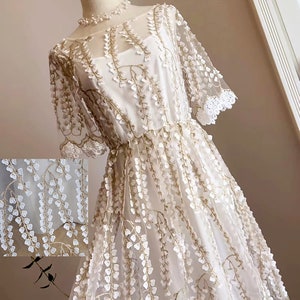 Pode incluir: Um vestido branco com uma sobreposição transparente e um bordado floral delicado. O vestido tem um decote redondo e mangas curtas.