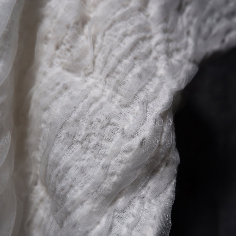 May include: White lace fabric with a textured, ruffled design. The fabric is soft and delicate, with a subtle sheen.