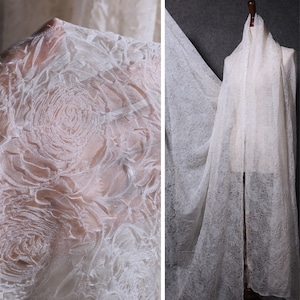 Könnte beinhalten: Off-white floral Spitze Stoff. Der Stoff hat ein zartes, transparentes Aussehen mit einem erhabenen Rosenmuster. Der Stoff ist über eine Schaufensterpuppe drapiert und zeigt seine Transparenz und Textur.