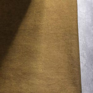 K&ouml;nnte beinhalten: Nahaufnahme eines rechteckigen, goldbraunen Stoffes mit subtiler Textur. Der Stoff ist gefaltet und zeigt auf einer Seite einen dunkleren Schatten. Der Hintergrund ist eine hellgraue, strukturierte Oberfl&auml;che.