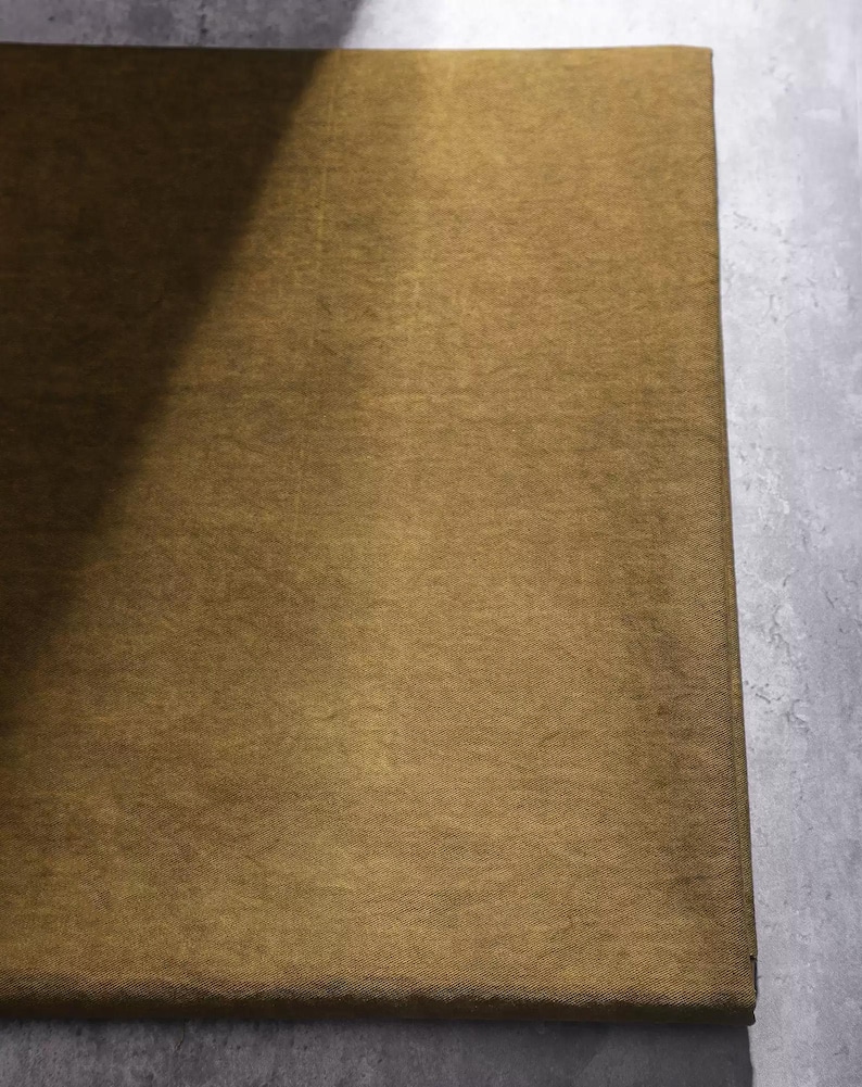 K&ouml;nnte beinhalten: Nahaufnahme eines rechteckigen, goldbraunen Stoffes mit subtiler Textur. Der Stoff ist gefaltet und zeigt auf einer Seite einen dunkleren Schatten. Der Hintergrund ist eine hellgraue, strukturierte Oberfl&auml;che.