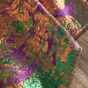 May include: Close-up of a shimmering fabric with a textured, abstract design. The fabric features a blend of colors, including green, purple, and bronze, creating a dynamic and visually rich pattern. The material appears to be folded, revealing its layered texture.