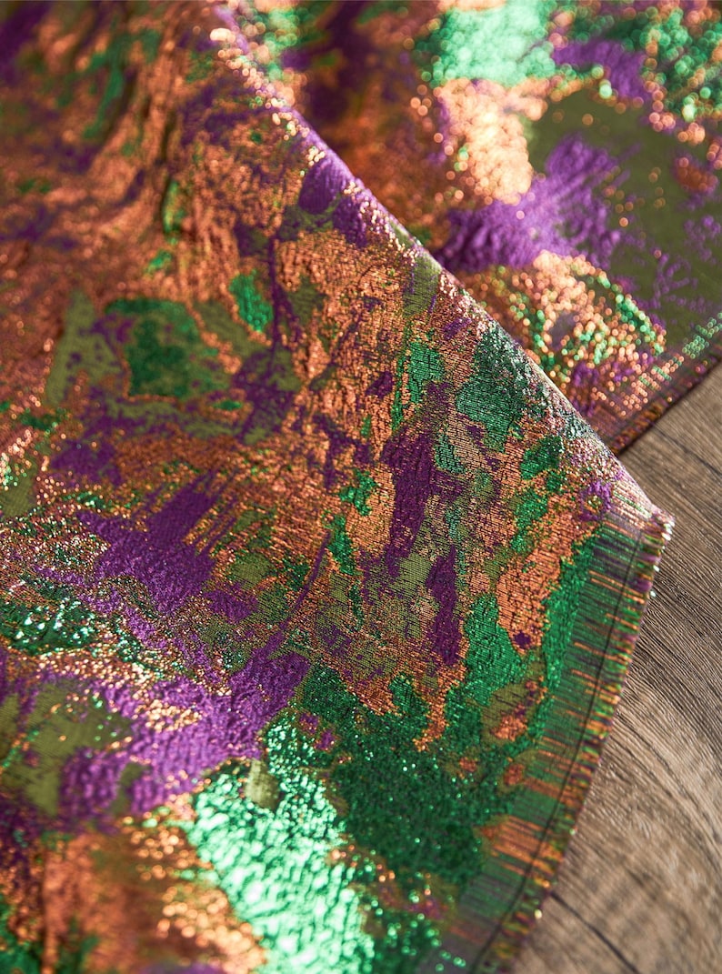May include: Close-up of a shimmering fabric with a textured, abstract design. The fabric features a blend of colors, including green, purple, and bronze, creating a dynamic and visually rich pattern. The material appears to be folded, revealing its layered texture.