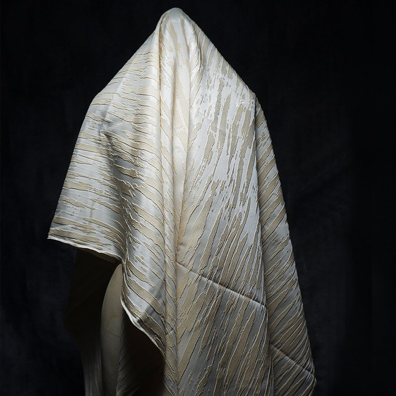 May include: A white and gold patterned fabric draped over a mannequin. The fabric has a subtle sheen and a textured surface.