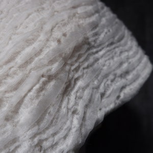 May include: White lace fabric with a ruffled texture. The fabric is soft and delicate, with a subtle sheen.