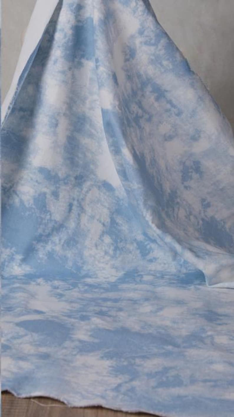 Op de afbeelding: Een lichtblauwe stof met een wit tie-dye patroon. De stof is gevouwen en gedrapeerd over een oppervlak.