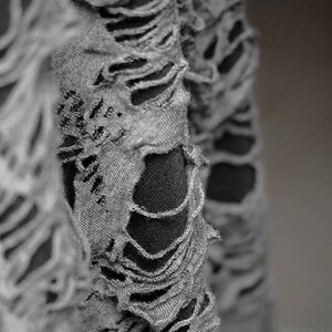 May include: Close-up of a gray fabric with a distressed, ripped look. The fabric has many holes and tears, creating a textured and edgy appearance.