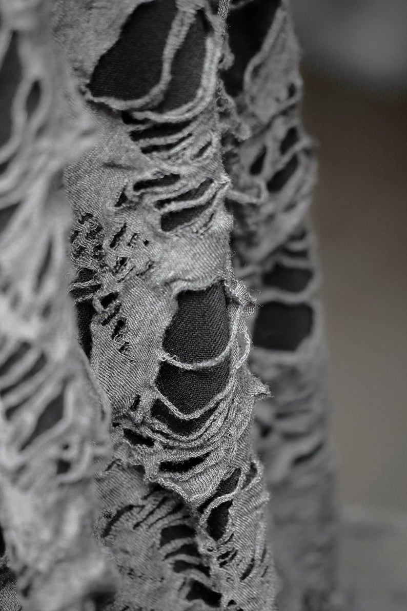 May include: Close-up of a gray fabric with a distressed, ripped look. The fabric has many holes and tears, creating a textured and edgy appearance.