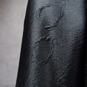May include: Close-up of a black fabric with a textured, embossed pattern. The fabric has a subtle sheen and a slightly wrinkled appearance.