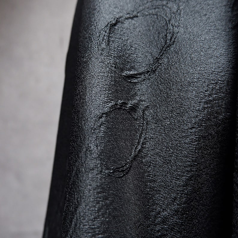May include: Close-up of a black fabric with a textured, embossed pattern. The fabric has a subtle sheen and a slightly wrinkled appearance.