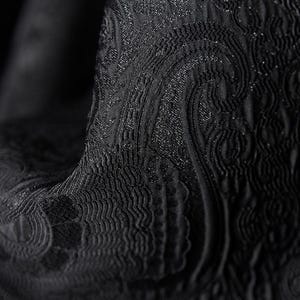 K&ouml;nnte beinhalten: Nahaufnahme eines schwarzen Stoffes mit einem strukturierten, wirbelnden Paisley-Muster. Der Stoff hat einen subtilen Glanz, mit Bereichen mit erhabener Textur und aufwendigen Details. Der Gesamteffekt ist elegant und luxuri&ouml;s und deutet auf ein hochwertiges Textil hin.