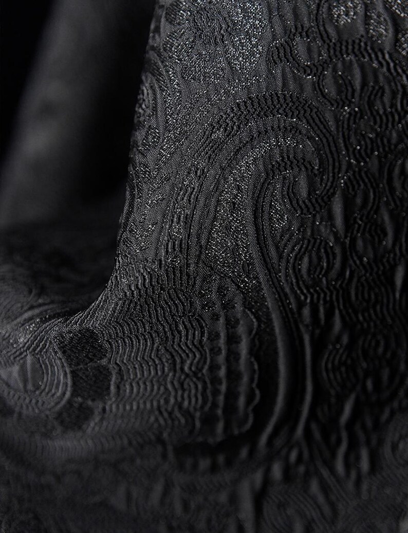 K&ouml;nnte beinhalten: Nahaufnahme eines schwarzen Stoffes mit einem strukturierten, wirbelnden Paisley-Muster. Der Stoff hat einen subtilen Glanz, mit Bereichen mit erhabener Textur und aufwendigen Details. Der Gesamteffekt ist elegant und luxuri&ouml;s und deutet auf ein hochwertiges Textil hin.