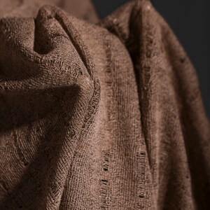 May include: Close-up of a brown fabric with a textured weave. The fabric is folded and draped, creating soft folds and shadows.