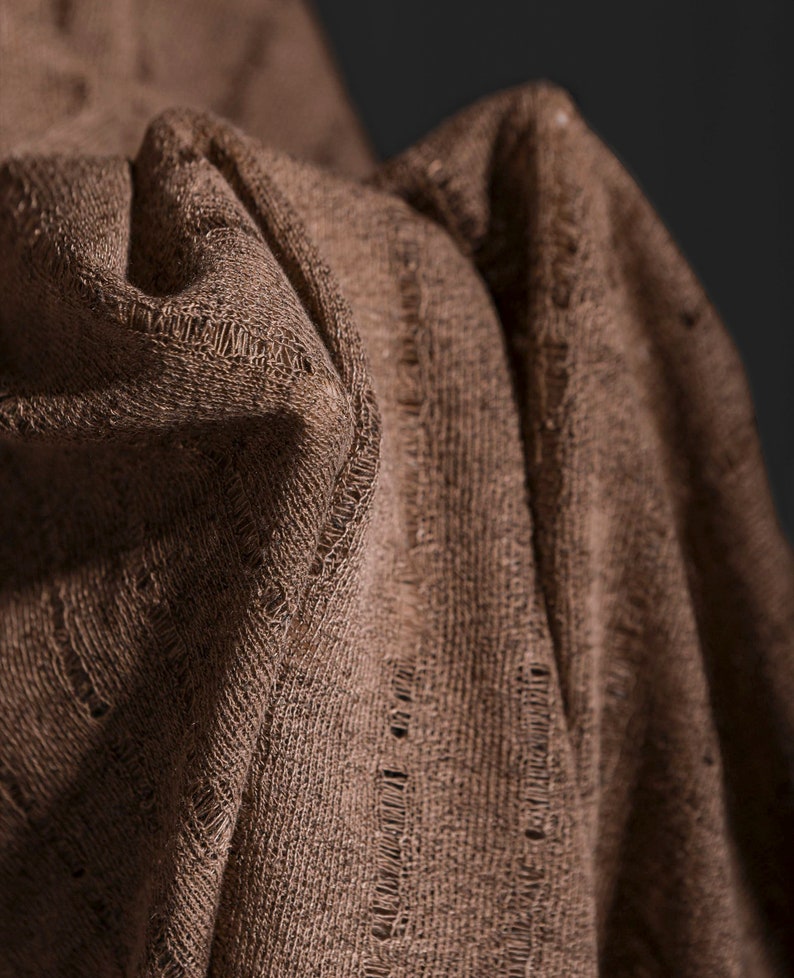 May include: Close-up of a brown fabric with a textured weave. The fabric is folded and draped, creating soft folds and shadows.