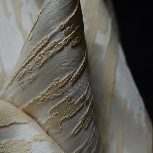 May include: Close-up of a white fabric with a subtle, embossed pattern of gold lines and shapes.