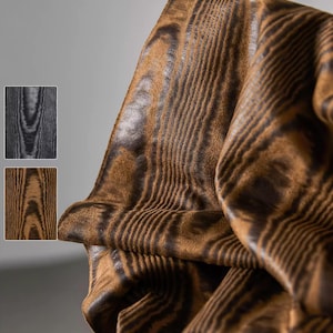 Op de afbeelding: Close-up van een bruin en zwart patroonstof met een houtnerf ontwerp. De stof is gedrapeerd en toont plooien en textuur. Twee kleinere stalen van hetzelfde patroon in grijs en bruin staan op de achtergrond.