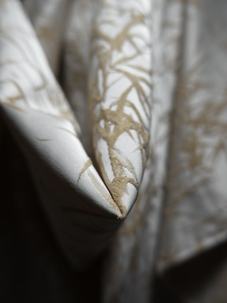 May include: Close-up of a white fabric with a subtle, embossed pattern of brown branches. The fabric is folded in a V-shape, creating a sharp crease.