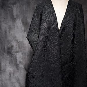 K&ouml;nnte beinhalten: Schwarze Jacke mit V-Ausschnitt und gepr&auml;gtem Blumenmuster. Das Kleidungsst&uuml;ck wird auf einer Schaufensterpuppe pr&auml;sentiert. Der Hintergrund ist eine dunkle, strukturierte Oberfl&auml;che.