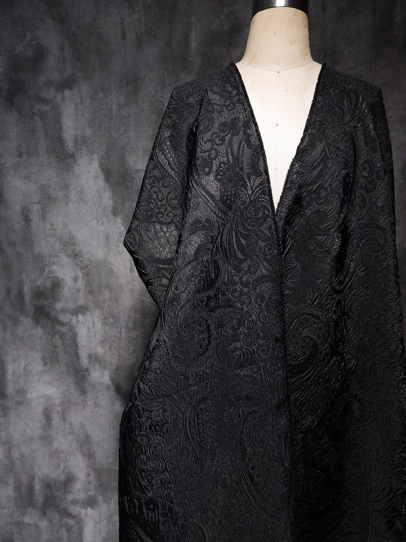 K&ouml;nnte beinhalten: Schwarze Jacke mit V-Ausschnitt und gepr&auml;gtem Blumenmuster. Das Kleidungsst&uuml;ck wird auf einer Schaufensterpuppe pr&auml;sentiert. Der Hintergrund ist eine dunkle, strukturierte Oberfl&auml;che.