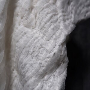 May include: White lace fabric with a delicate, textured pattern. The fabric is gathered and ruffled, creating a soft and flowing look.