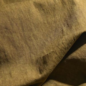 K&ouml;nnte beinhalten: Nahaufnahme eines olivgr&uuml;nen Stoffes mit diagonal gerippter Textur. Der Stoff ist gefaltet, wodurch Schatten und Farbvariationen entstehen. Das Material scheint weich zu sein und m&ouml;glicherweise f&uuml;r Kleidung oder Heimdekoration verwendet zu werden.