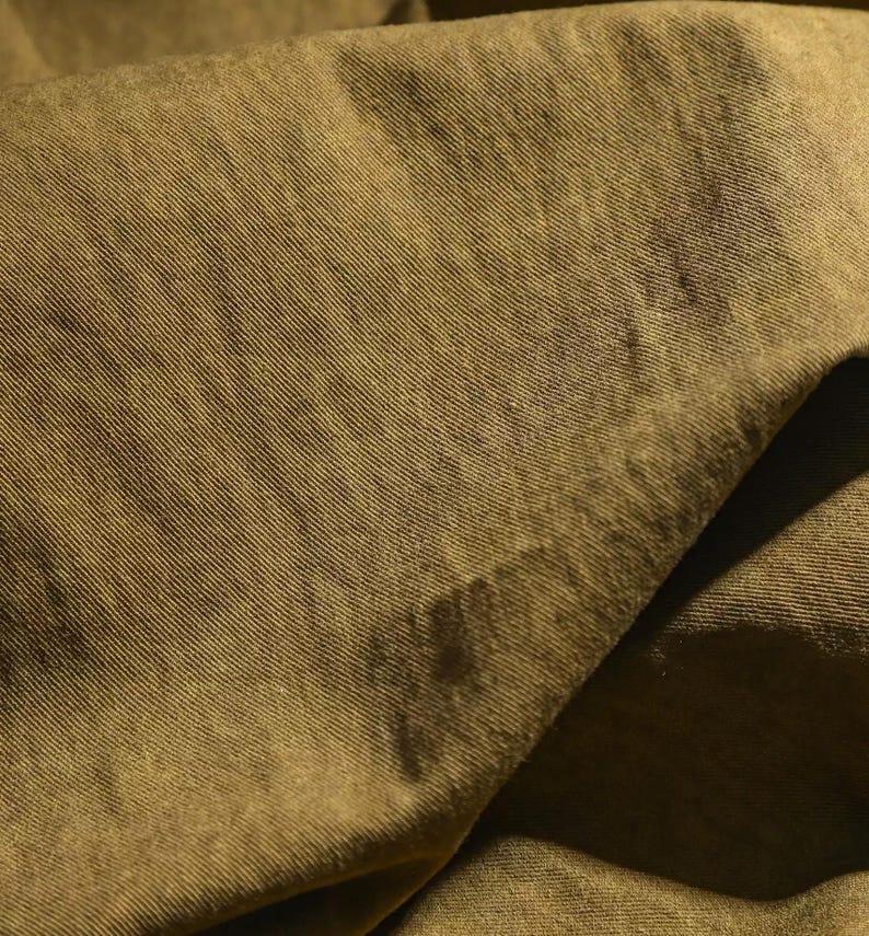 K&ouml;nnte beinhalten: Nahaufnahme eines olivgr&uuml;nen Stoffes mit diagonal gerippter Textur. Der Stoff ist gefaltet, wodurch Schatten und Farbvariationen entstehen. Das Material scheint weich zu sein und m&ouml;glicherweise f&uuml;r Kleidung oder Heimdekoration verwendet zu werden.