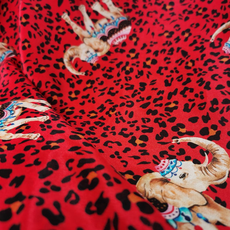 May include: Red fabric with a repeating pattern of black leopard spots and colorful elephants wearing ornate headbands.