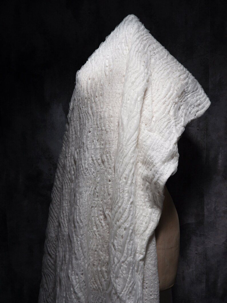 May include: White fabric with a textured, wavy pattern. The fabric is draped over a form and has a delicate, airy feel.