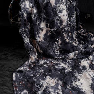 May include: A draped fabric with a tie-dye pattern in shades of black, white, and tan. The fabric has a textured, fringed appearance, with frayed edges and a layered design. The material is likely used for clothing or home decor.