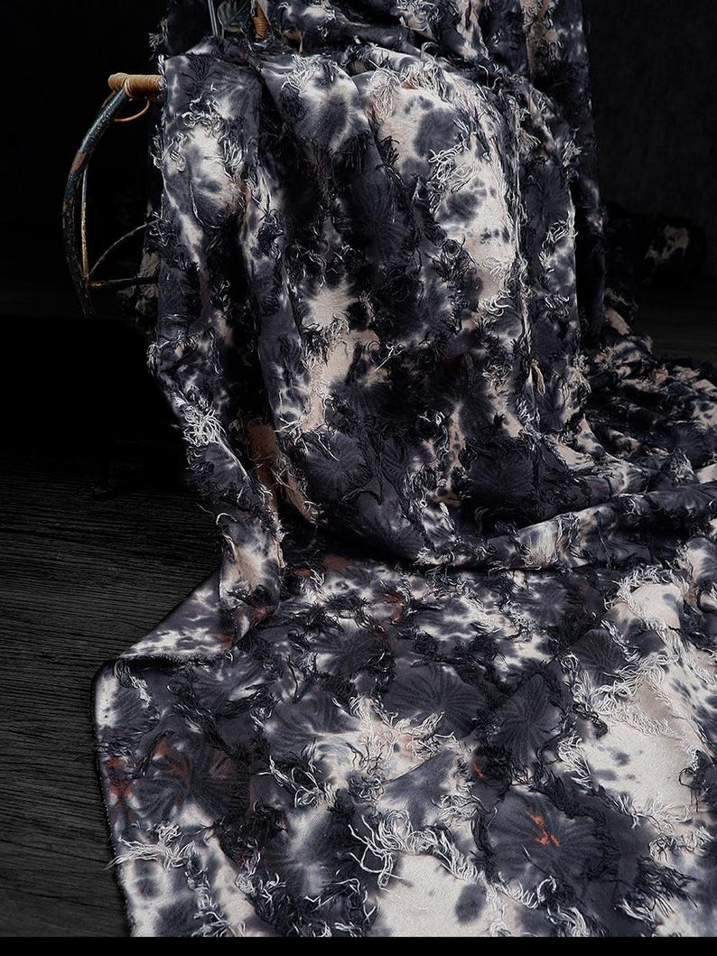 May include: A draped fabric with a tie-dye pattern in shades of black, white, and tan. The fabric has a textured, fringed appearance, with frayed edges and a layered design. The material is likely used for clothing or home decor.
