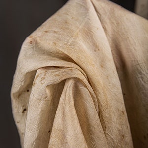 May include: A close-up of a white fabric with a textured pattern and brown stains. The fabric is folded and draped, creating soft folds and wrinkles.