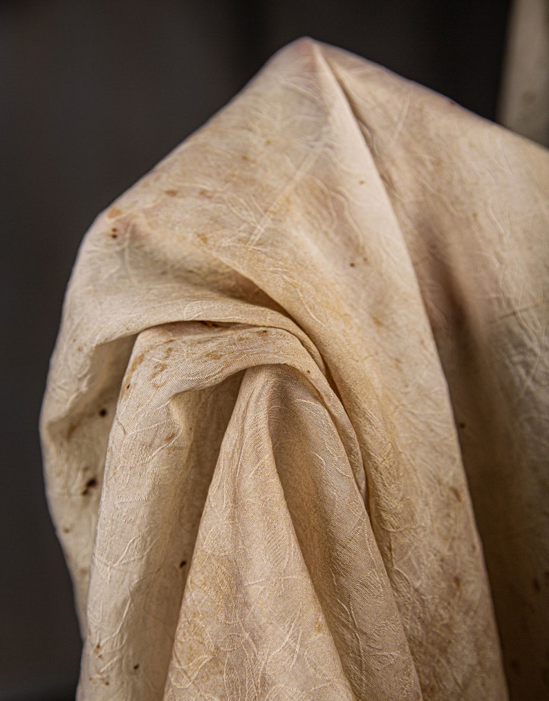 May include: A close-up of a white fabric with a textured pattern and brown stains. The fabric is folded and draped, creating soft folds and wrinkles.