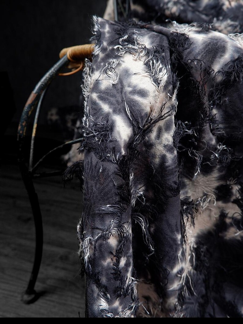 May include: Close-up of a tie-dye fabric with a distressed, fringed edge. The fabric features a pattern of black and white swirls, creating a textured appearance. The fabric is draped over a chair.