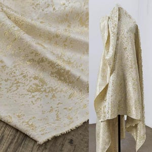 以下が含まれることがあります： 金色のテクスチャパターンが施されたクリーム色の生地。生地は型にドレープされ、平らに置かれ、詳細なデザインが示されています。素材は微妙な光沢があり、高級感を高めています。生地の端が見え、その生の未完成の状態が明らかになっています。