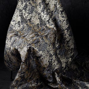 Puede incluir: Tela texturizada con un patrón abstracto en negro, gris y dorado. El material está drapeado sobre una silla, resaltando su forma fluida y su diseño intrincado. La paleta de colores crea una estética lujosa y artística.