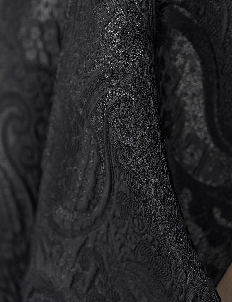 K&ouml;nnte beinhalten: Nahaufnahme eines schwarzen Stoffes mit detailliertem Paisley-Muster. Der Stoff hat eine strukturierte Oberfl&auml;che mit dezentem Glanz, der Tiefe und visuelles Interesse erzeugt. Das Design zeigt wirbelnde, geschwungene Motive.
