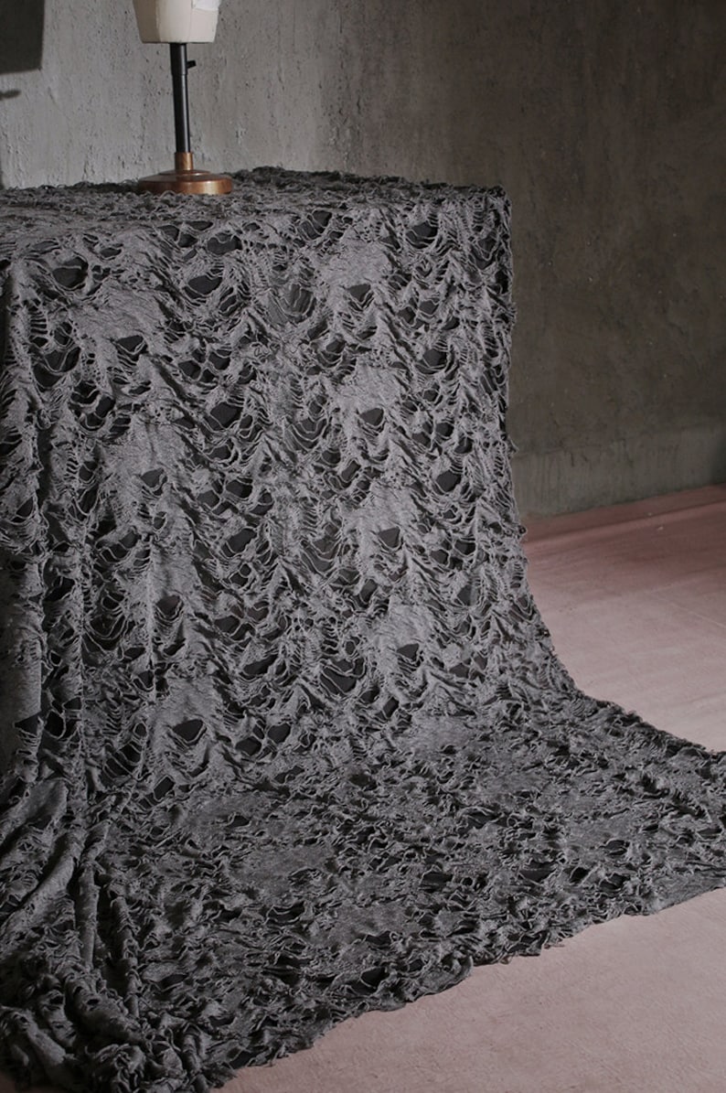 May include: Gray distressed fabric with a textured, ripped, and torn pattern. The fabric is draped over a surface and appears to be a backdrop or stage prop.