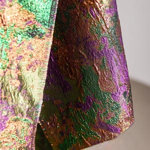 May include: Close-up of a textured fabric with a shimmering, iridescent effect. The material features a blend of colors, including green, purple, and gold, creating a dynamic and visually rich appearance. The fabric appears to be part of a garment.