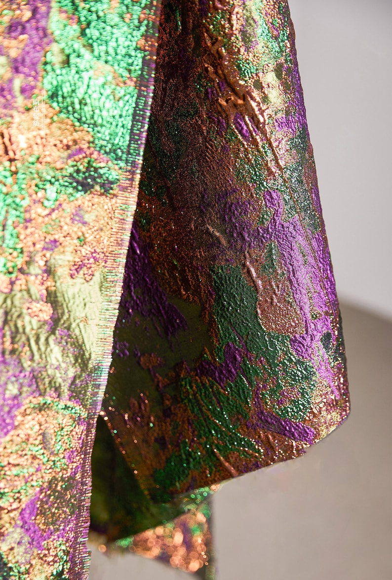 May include: Close-up of a textured fabric with a shimmering, iridescent effect. The material features a blend of colors, including green, purple, and gold, creating a dynamic and visually rich appearance. The fabric appears to be part of a garment.