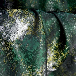 May include: Close-up of a fabric with a textured, abstract design. The fabric features a dark green base with splashes of silver, yellow, and lighter green, creating a mottled effect. The fabric is folded, showing depth and dimension.